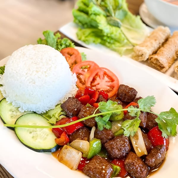 Vietnamese shaking beef (Bo Luc Lac) with rice, tomatoes, cucumbers, and spring rolls in the background.